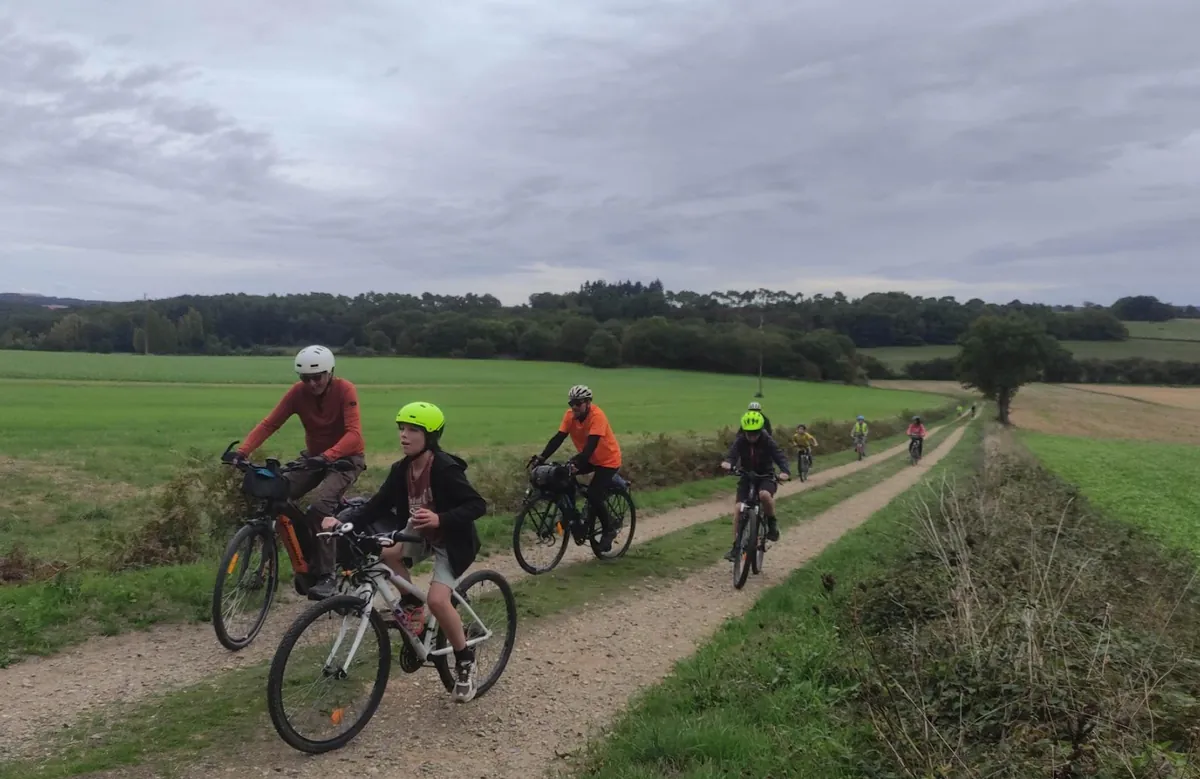 Quatre cyclistes roulent sur un chemin de terre entre les champs, sous un ciel gris